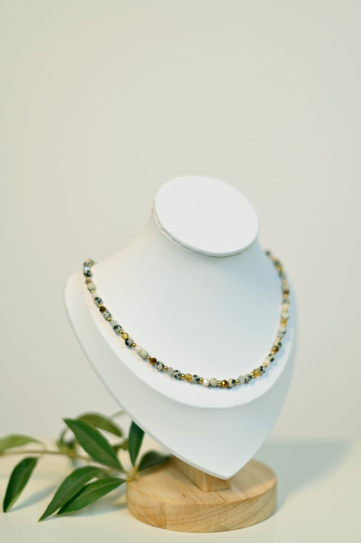 Perlenkette auf einem Kettenaufsteller mit braunen und goldenen Perlen und Perlen aus Dalmatiner-Jaspis. Im Hintergrund sieht man einen Olivenzweig 